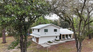 A two-story white house with a metal roof, surrounded by trees and a small porch with chairs.