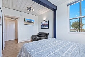 A bedroom with a bed, a black leather chair, a framed picture on the wall, and a window with a view of buildings and trees.