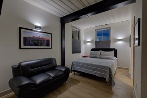A bedroom with a bed, a black leather sofa, a framed picture on the wall, and a window with blinds.