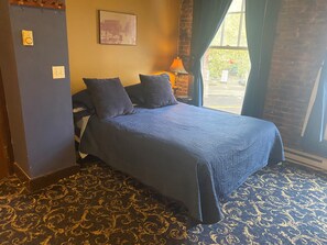 Une chambre à coucher avec un lit, une literie bleu foncé, une moquette à motifs, une table de chevet avec une lampe et une fenêtre avec des rideaux.