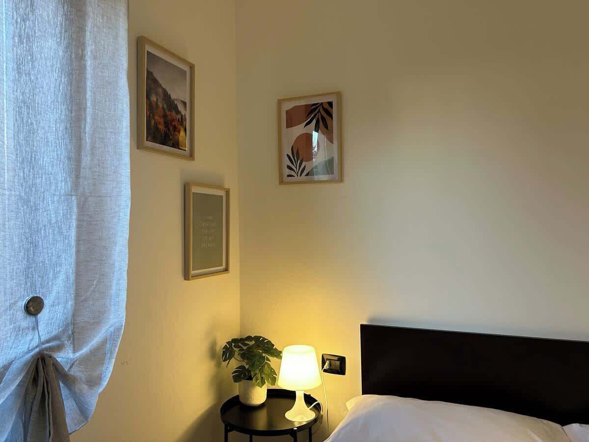 Habitación de hotel con cama, mesita de noche con lámpara, planta en maceta y dos cuadros en la pared.