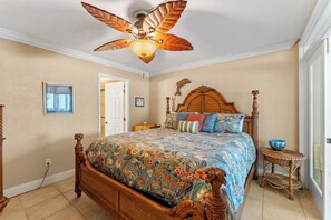 A bedroom with a wooden bed, a ceiling fan, a small table, and a door leading to another room. - Oceanfront Escape at Smyrna Beach Club -601 (New Smyrna Beach)