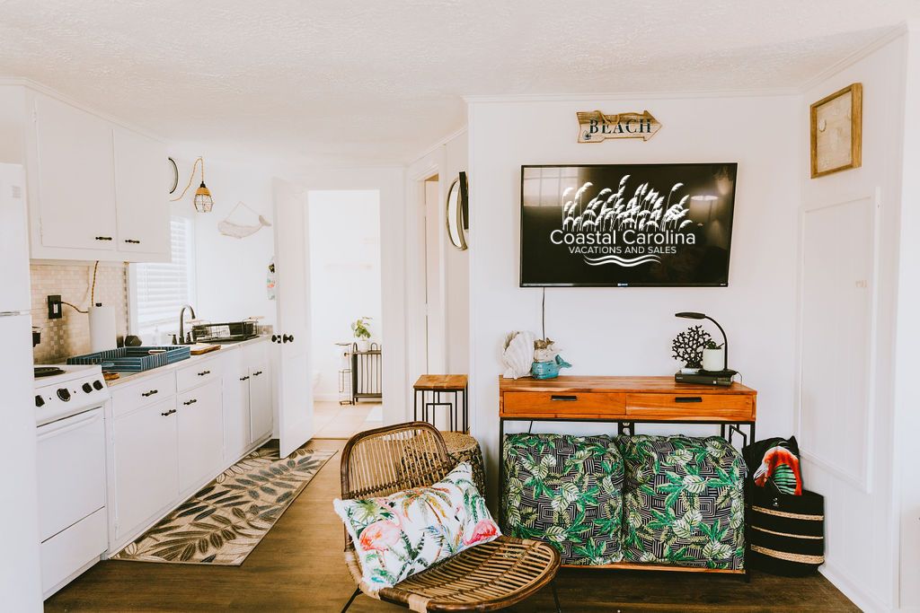 A kitchen with white cabinets, a dining area with a wicker chair and patterned ottoman, and a TV displaying 'Coastal Carolina Vacations and Sales'.