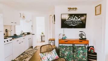 A kitchen with white cabinets, a dining area with a wicker chair and patterned ottoman, and a TV displaying 'Coastal Carolina Vacations and Sales'.
