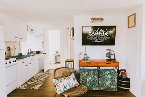 A kitchen with white cabinets, a dining area with a wicker chair and patterned ottoman, and a TV displaying 'Coastal Carolina Vacations and Sales'.