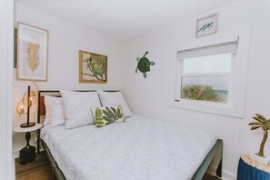 A bedroom with a bed, a bedside table with a lamp, and wall art including a bird, a turtle, and a plant.
