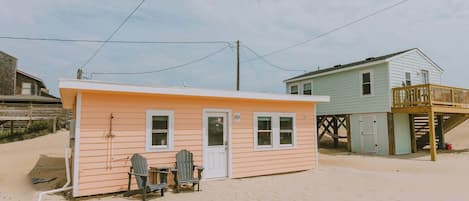 A small beach house with a single door and two windows, a deck, and a chair outside.