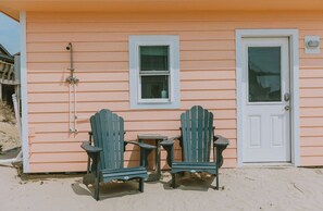 Ein Strandhaus mit zwei Adirondack-Stühlen, einem kleinen Tisch und einer Tür mit Fenster darüber.