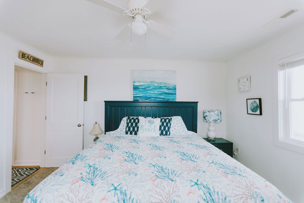 A bedroom with a bed, a ceiling fan, a painting of the ocean, a nightstand with a lamp, and a sign that says "BEACHHOUSE".