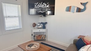 En stue med et fladskærms-tv på væggen, et træsofabord med en vævet kurv, et hvidt vedligeholdelsesbord med bøger og en sofa med stribede puder.