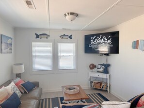 A living room with a sofa, a coffee table, a TV, and two windows with blinds. - Coastal Dune 3: Oceanfront Cottage on the Sand in Nags Head! (Nags Head)