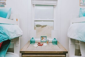 Una habitación con dos literas, una mesa de madera con una maqueta de una casa, un letrero que dice "STAY MAGICAL" y una ventana con vistas a dunas de arena.