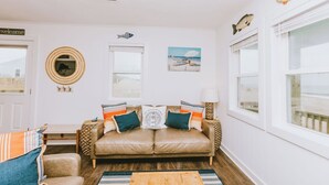 A cozy living room with a beige leather sofa, striped cushions, and a wooden coffee table. There are two windows with white frames, a wall-mounted fish decoration, and a welcome sign.