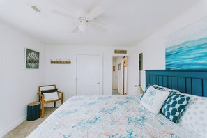 A bedroom with a bed, a ceiling fan, a chair, a door, and a hallway leading to another room. - Coastal Dune 3: Oceanfront Cottage on the Sand in Nags Head! (Nags Head)