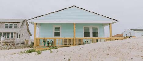 A beach house with a light blue exterior, a wooden deck, and a sloped roof.