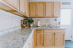 A kitchen with wooden cabinets, a granite countertop, a sink, and a 'welcome' sign. - Coastal Dune 3: Oceanfront Cottage on the Sand in Nags Head! (Nags Head)