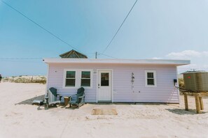 Una pequeña casa de playa de un piso con una puerta blanca, dos ventanas y un porche con una silla y una mesa.