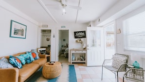Een goed verlichte woonkamer met een bruine leren bank, een rieten poef en een witte deur die naar een andere kamer leidt.
