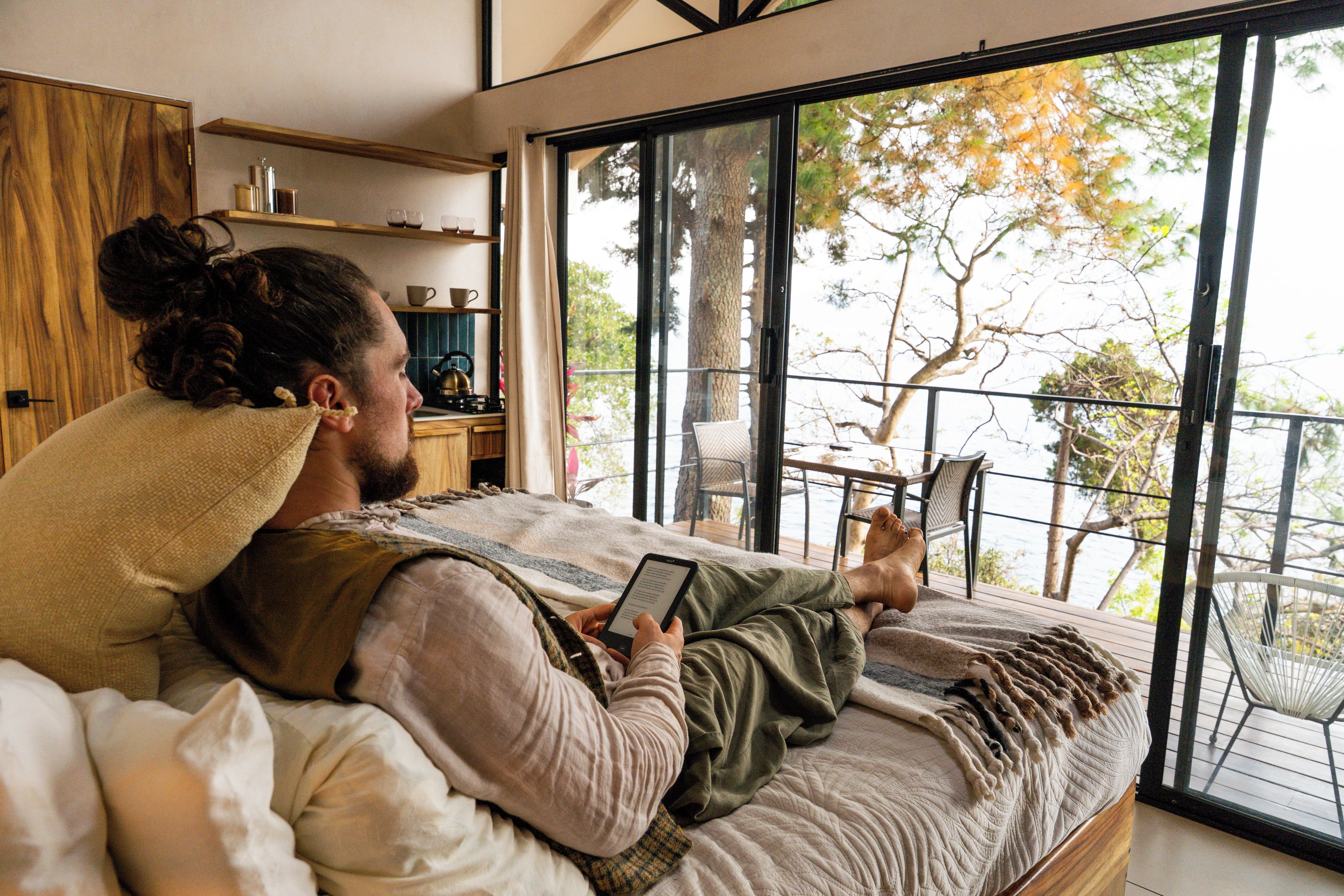 A person lying on a bed with a tablet, reading, in a room with a large window overlooking a balcony with outdoor furniture.