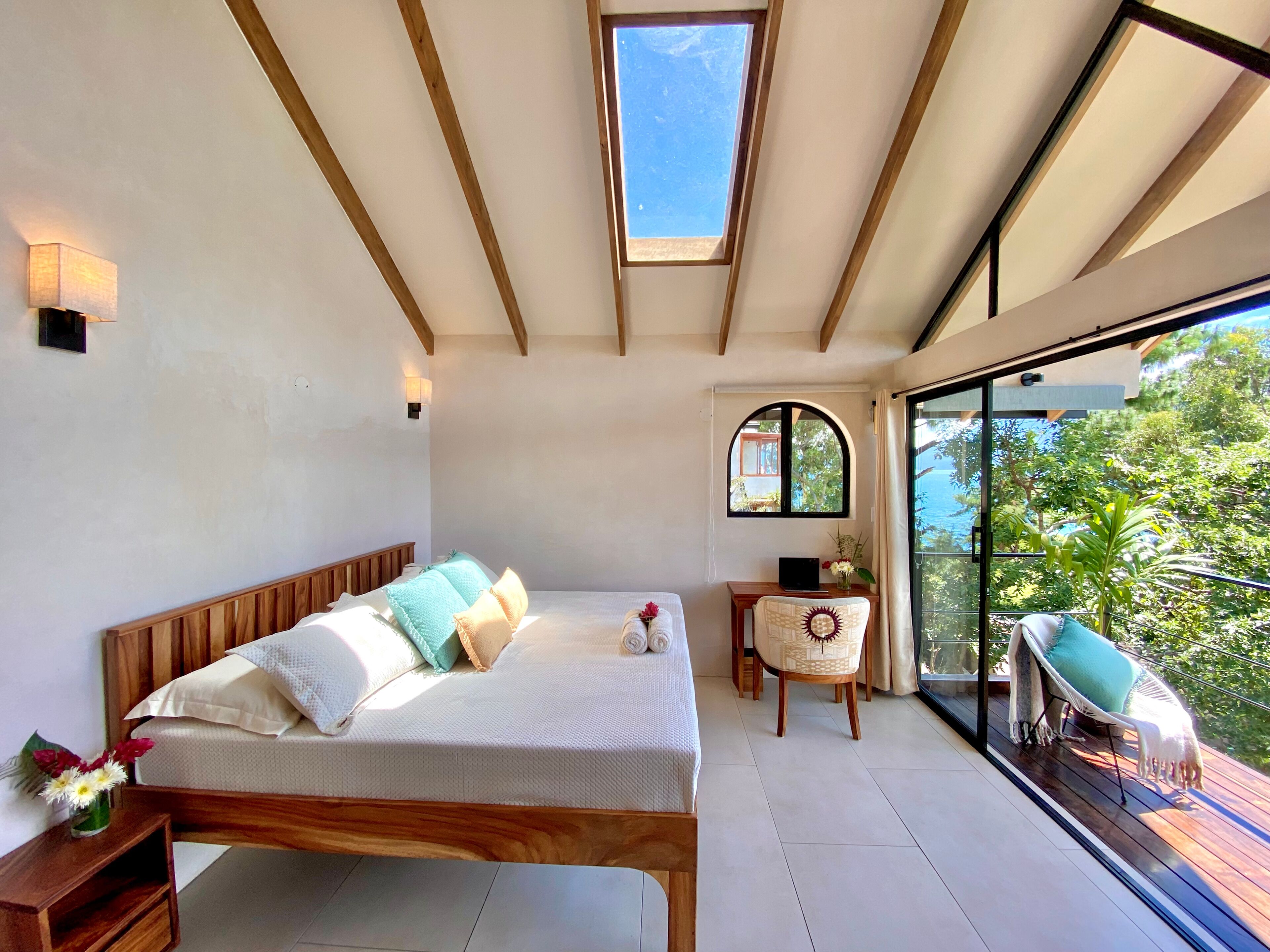 A bedroom with a wooden bed, a small desk, and a view of the outdoors through a large window.