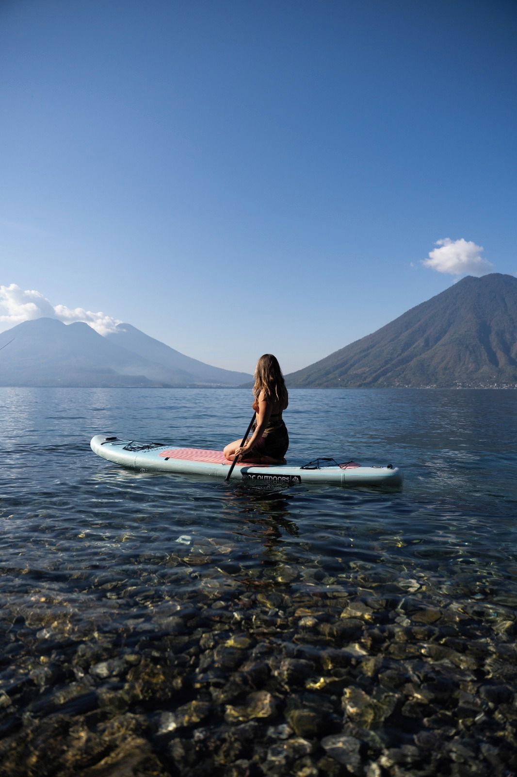 Una persona sentada en una tabla de remo en un cuerpo de agua, con montañas al fondo.