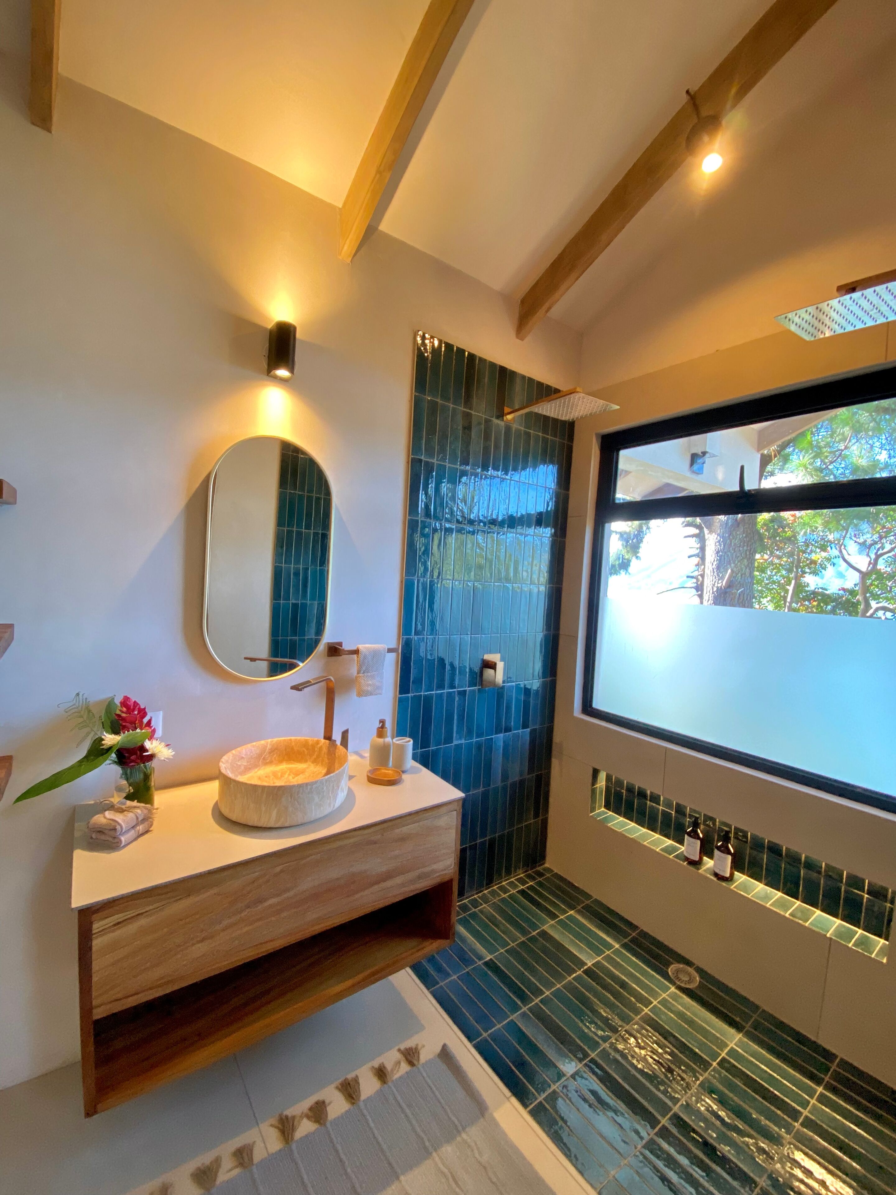Un baño con tocador de madera, un espejo redondo, zona de ducha con azulejos azules y una ventana con vistas a árboles.