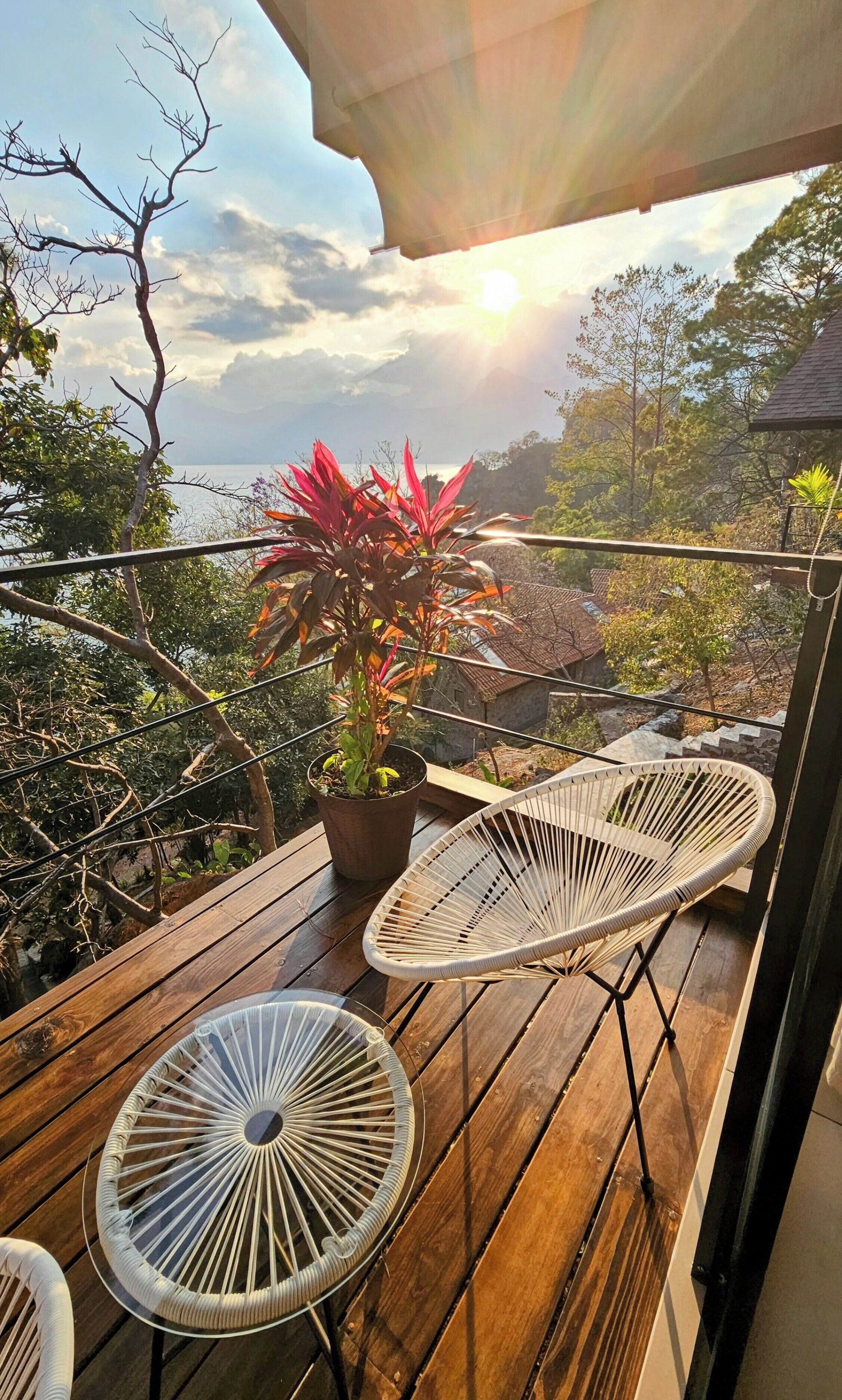 A wooden deck with two wicker chairs and a potted plant, overlooking a scenic landscape with trees and a view of the sea.