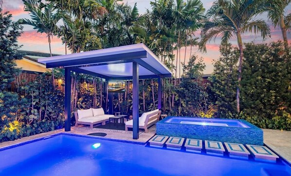 Zona de piscina con un área de estar cubierta, un jacuzzi y exuberante vegetación tropical.