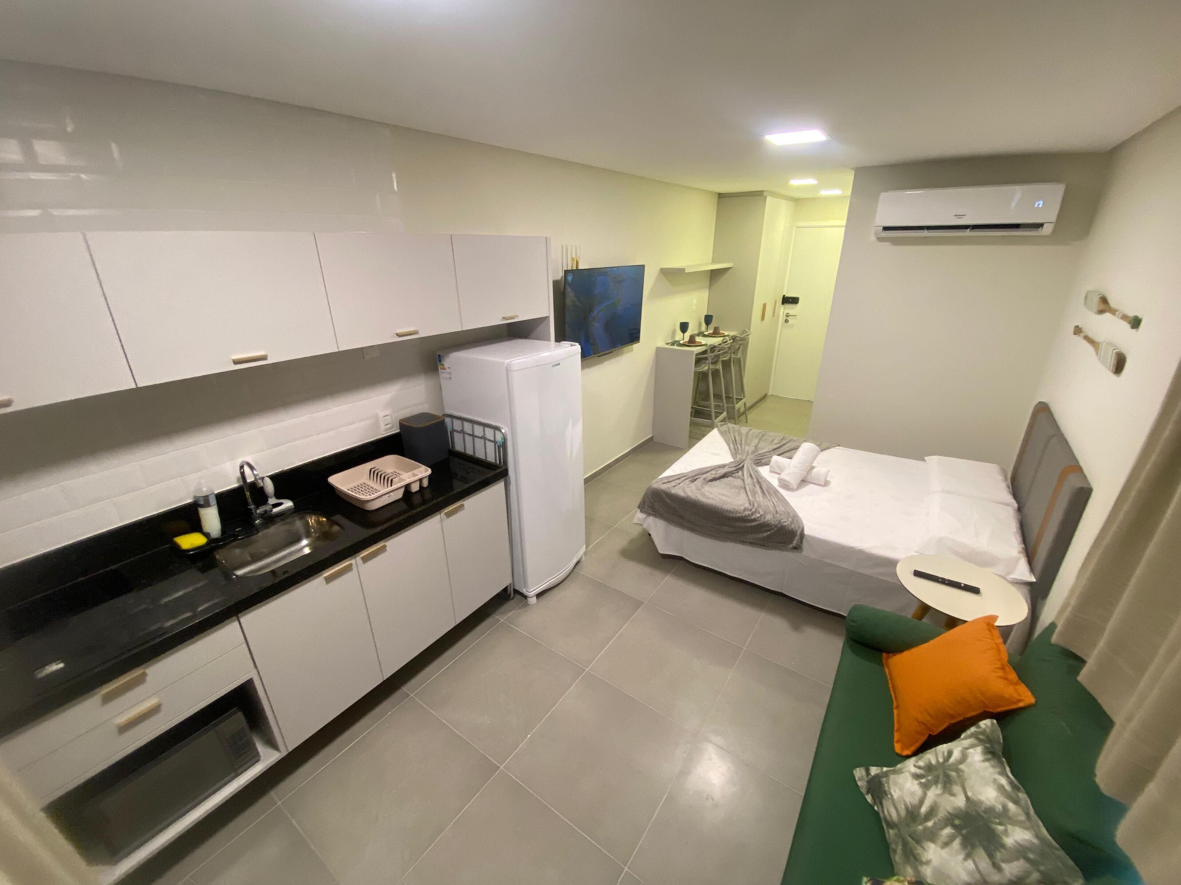 A compact hotel room with a kitchenette, a bed with a green and orange pillow, and a small seating area.