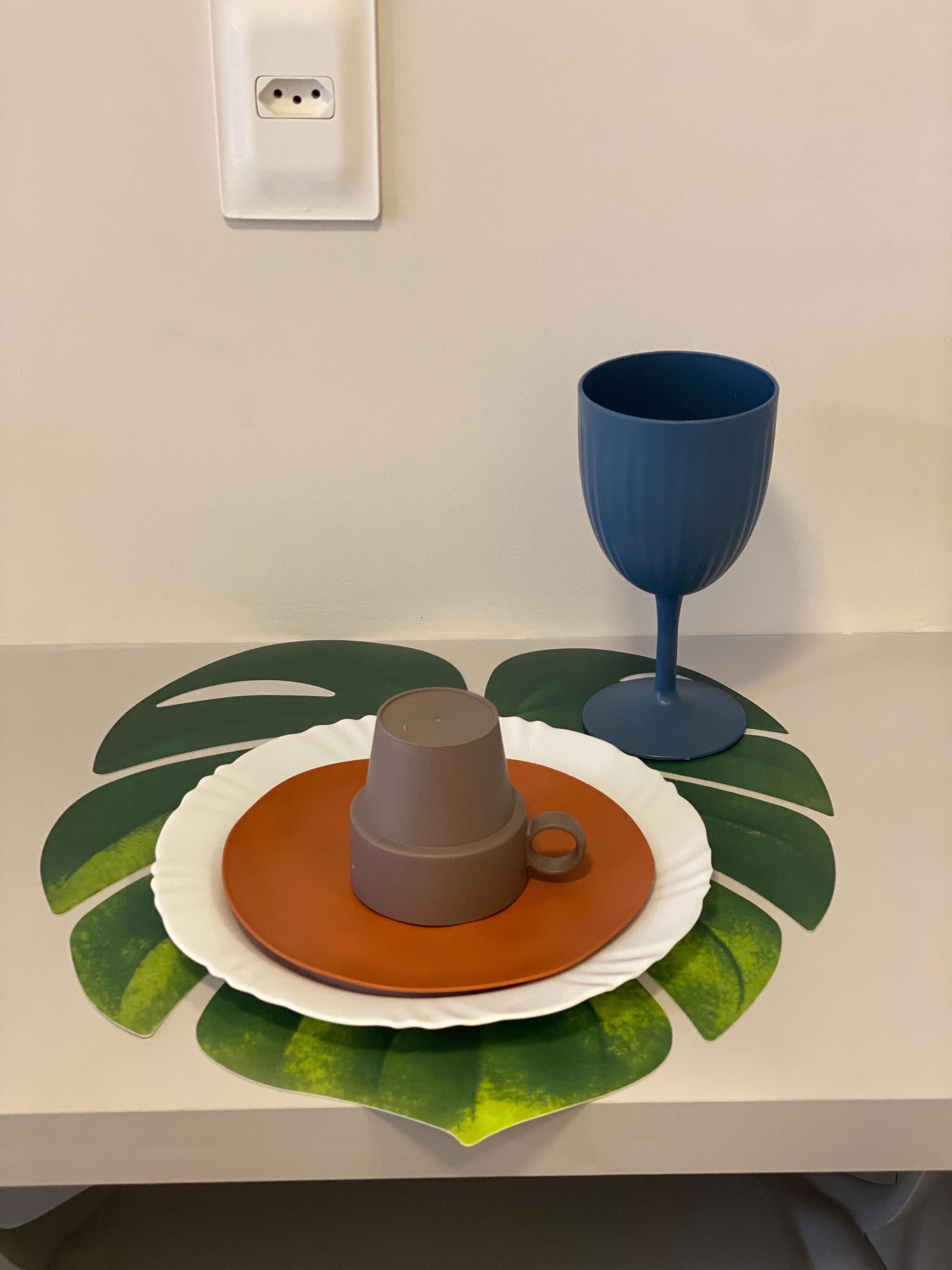 Uma mesa com uma caneca marrom sobre um prato laranja, uma chávena azul e um desenho de folha verde.