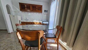 Uma área de refeições com mesa de vidro e cadeiras de madeira, uma cozinha com azulejos brancos e armários de madeira, e um chão ladrilhado.