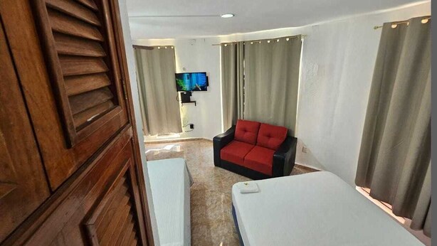 Quarto de hotel com porta de madeira, sofá vermelho e preto, cama e televisão na parede.
