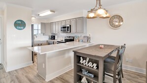 A modern kitchen with a central island, stainless steel appliances, and a wooden dining table with a 'relax' sign. - Beachside @ Seaside Beach & Racquet 2BR Condo • Sleeps 6 • Pools, Tennis & Beach Access (Orange Beach)