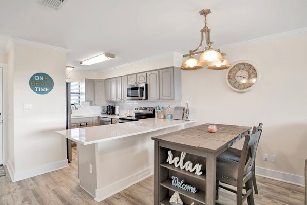 A modern kitchen with a central island, stainless steel appliances, and a wooden dining table with a 'relax' sign. - Beachside @ Seaside Beach & Racquet 2BR Condo • Sleeps 6 • Pools, Tennis & Beach Access (Orange Beach)