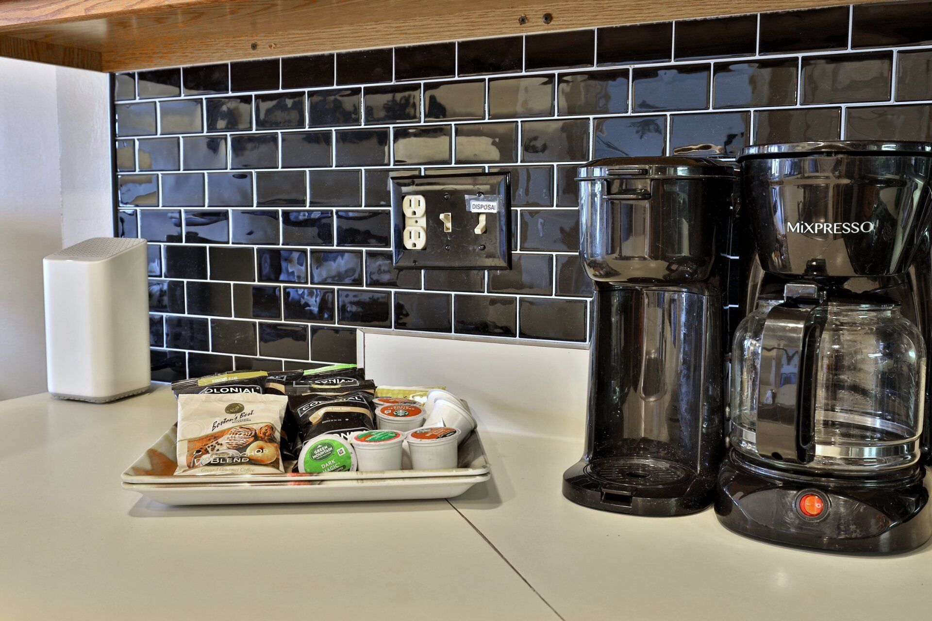 A kitchen with a black tiled backsplash, a coffee maker, and a plate of coffee pods.