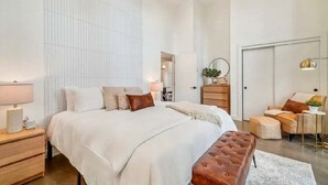 Une chambre moderne avec un grand lit, des tables de chevet, une commode et un coin salon comprenant un fauteuil et un pouf.