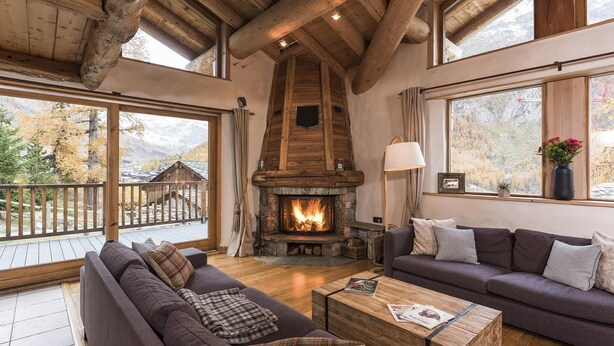 A cozy cabin interior with a stone fireplace, wooden beams, a balcony with a wooden railing, and large windows offering mountain views. - Navette Privative | Chalet cozy pour 10 + Jacuzzi (Val-d'Isère)