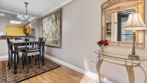 A dining area with a wooden table and chairs, a chandelier, a large framed painting, and a glass console table with a lamp and vase of flowers.