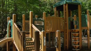 A wooden playground structure with slides and climbing elements, surrounded by trees.