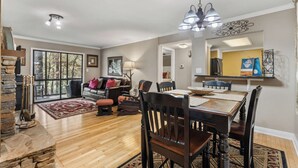 A spacious living room with a fireplace, dining area with wooden chairs, and a large window offering a view of trees.