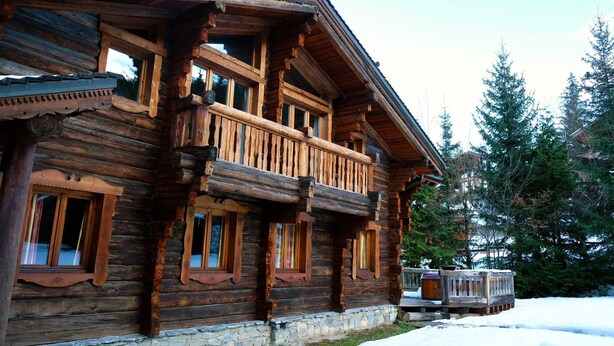 A log cabin with a balcony, stone foundation, and surrounded by snow and trees.