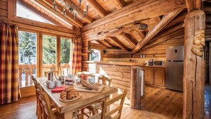 Interior rústico de una cabaña con una mesa de madera dispuesta para el desayuno, una cocina con armarios de madera y un ventanal con vistas a un bosque.