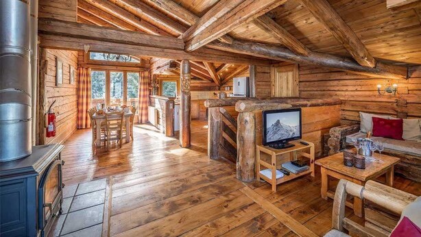 Interior de una cabaña de troncos con comedor, chimenea, televisor y una zona de estar acogedora.