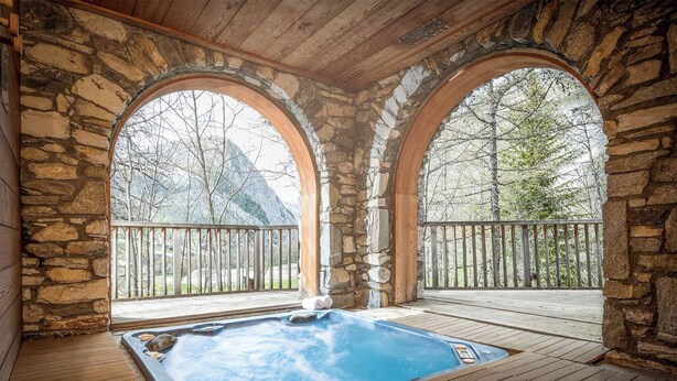 Een hottub met stenen muren, een houten terras en uitzicht op bomen en bergen.