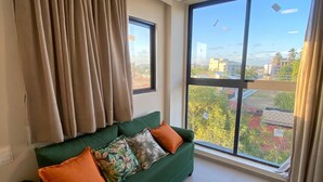 Un sofá verde con cojines naranjas, una ventana con vistas a edificios y árboles, y una lámpara en el techo.