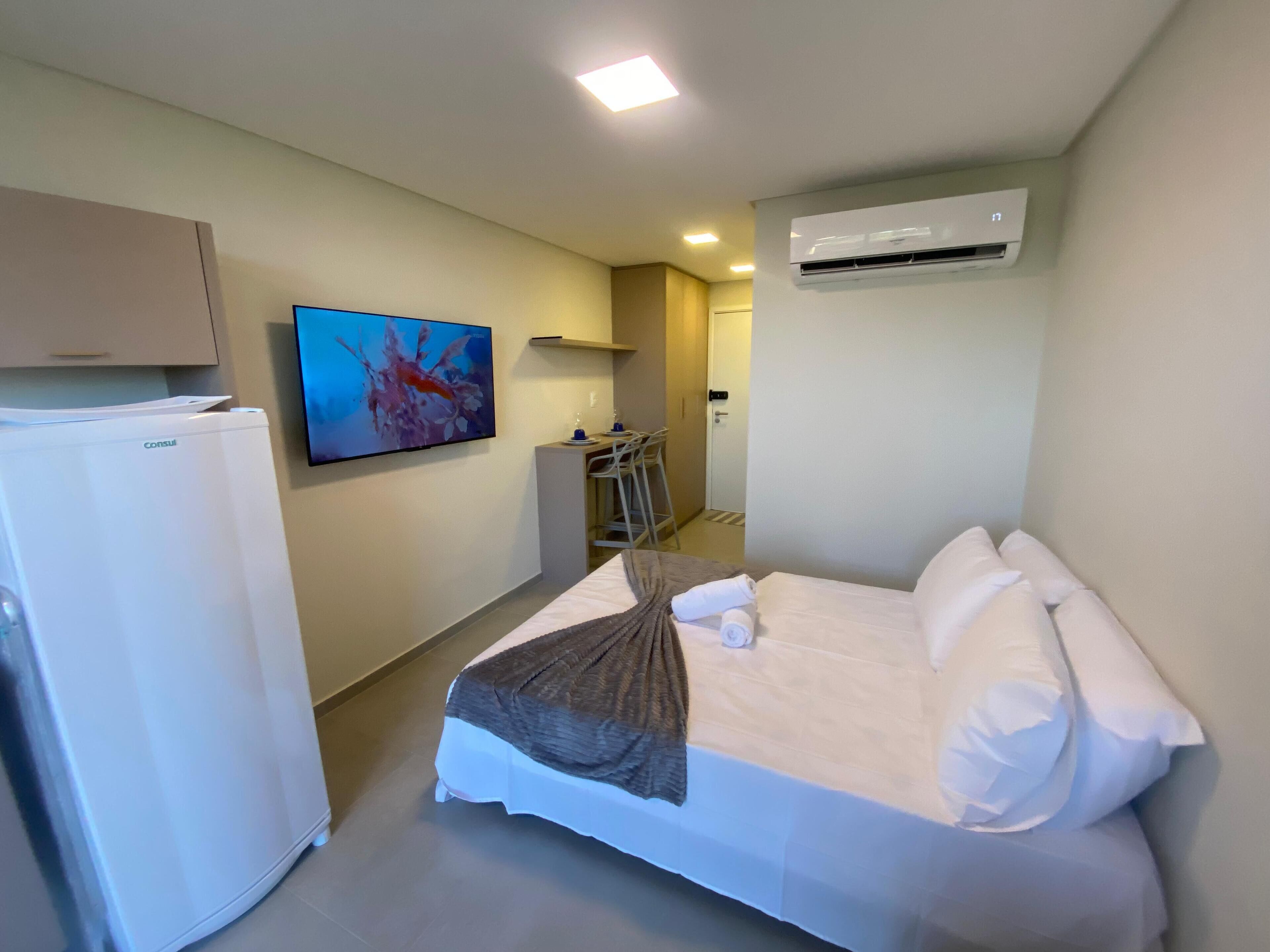 A hotel room with a bed, a desk, a chair, a television, an air conditioner, and a refrigerator.