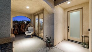 A covered patio with a cushioned chair, a potted plant, and a glass door. - Casa Desert Muse: Heated Pool, Spa, Fire, Golf, Games, Pool Table, Art!

 (Indio)