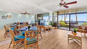 A spacious dining area with wooden chairs and a table set for a meal, a television, and a balcony with a view of the ocean. - Sun-Kissed Retreat with Oceanfront Views (Lahaina)