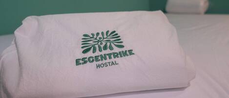 Una cama bien hecha con sábana blanca y una toalla doblada encima. La toalla tiene un logotipo y el texto "ESCENTRIKE HOSTAL".