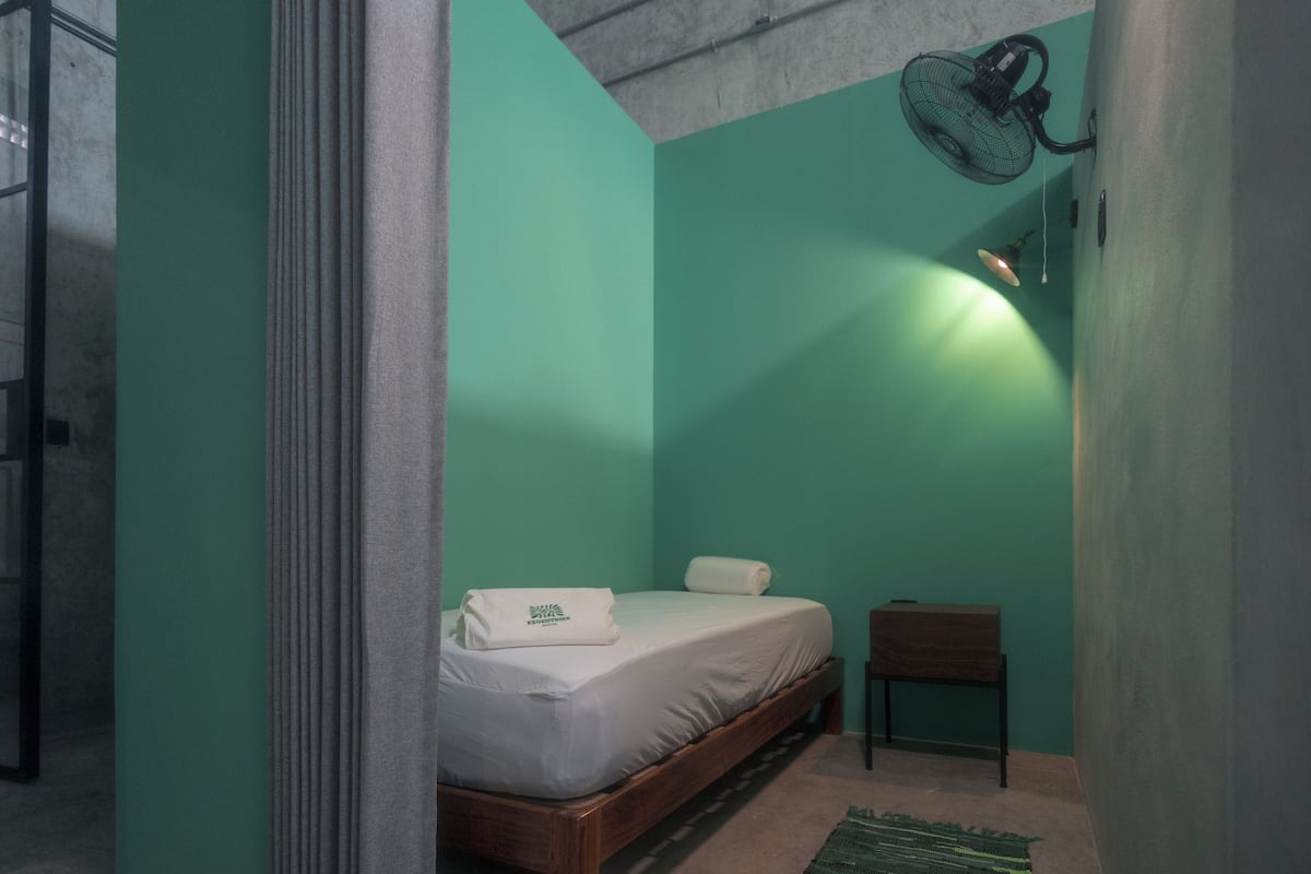 A small, cozy room with a single bed, a chair, and a standing fan.